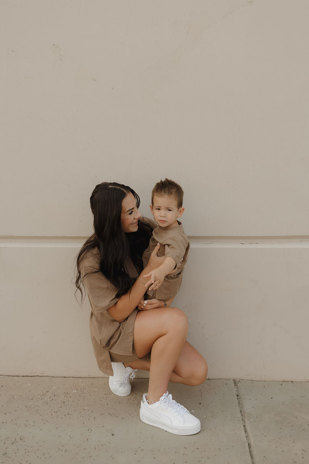 A woman with long dark hair kneels by a beige wall, holding a young child dressed in the forever french baby Child Short Set | Patina. Both wear matching cozy biker shorts and oversized tees as they share a sweet moment together.