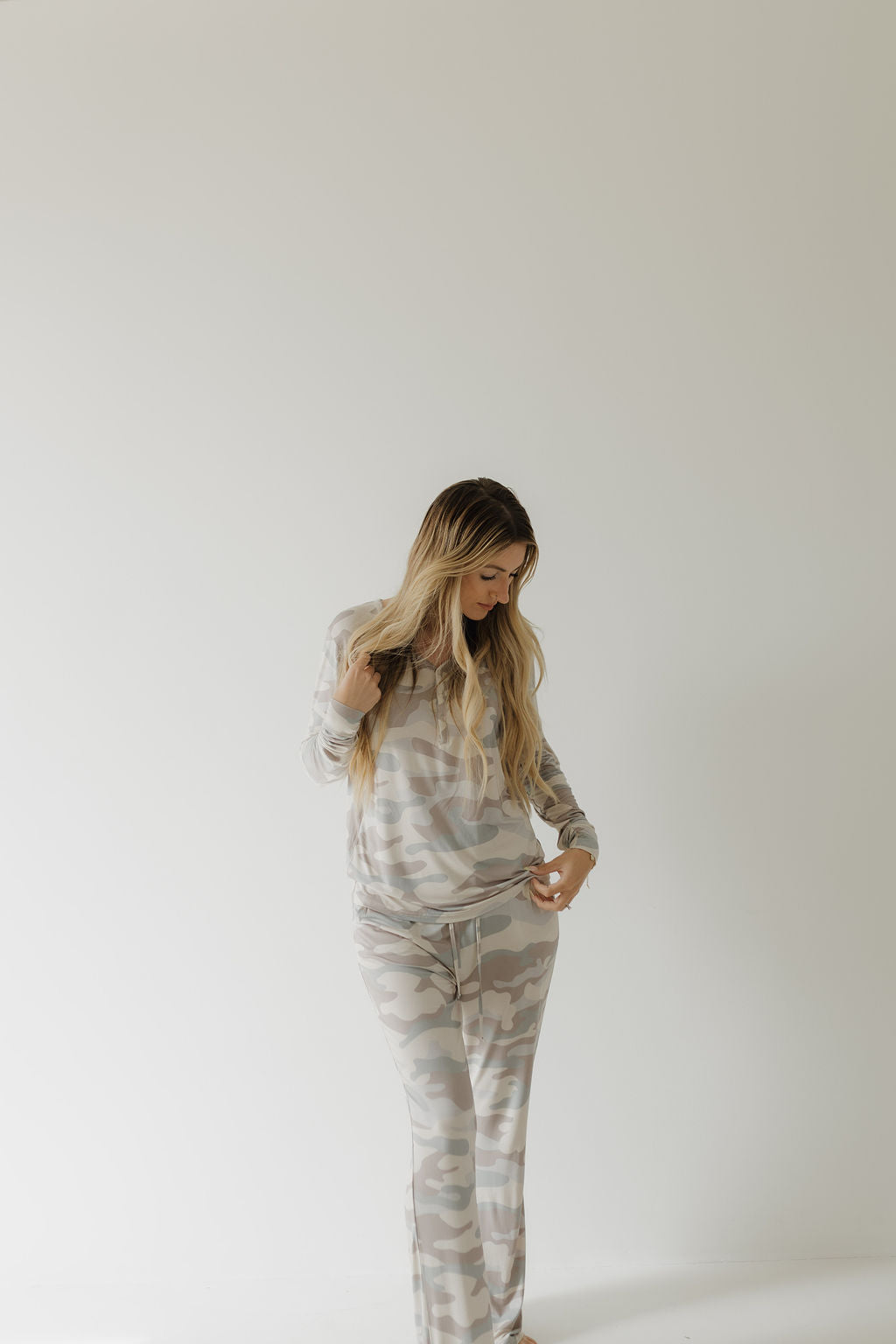 A person with long blonde hair wears the Women's Bamboo Pajama in camouflage by forever french baby. Against a plain white background, they look downward while slightly adjusting their hypoallergenic gray and white outfit.