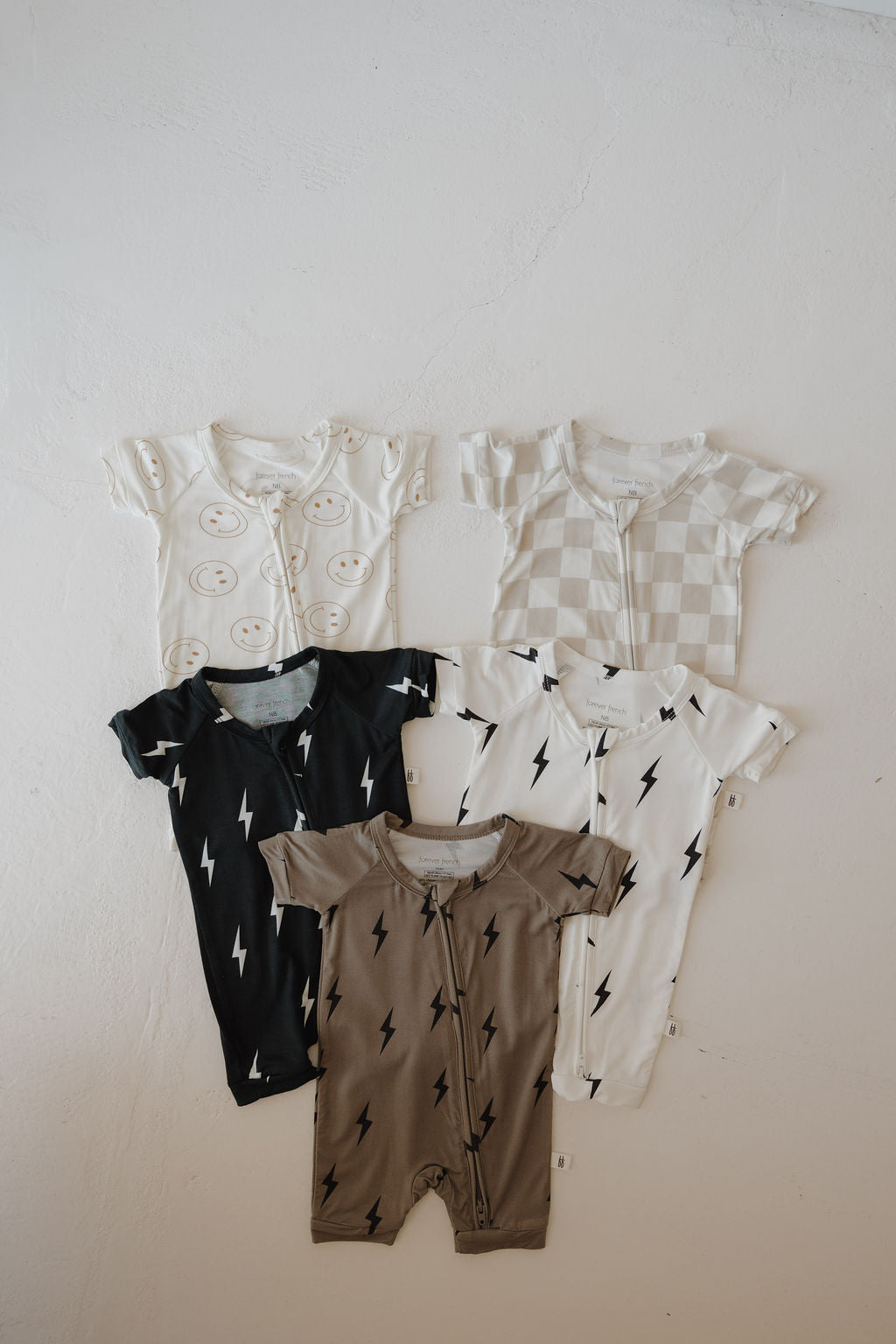 Five Bamboo Zip Shortie onesies by forever french baby are displayed on a white surface, featuring smiley faces, checkerboard, and lightning bolt designs. These breathable options keep your little one stylish and comfortable.