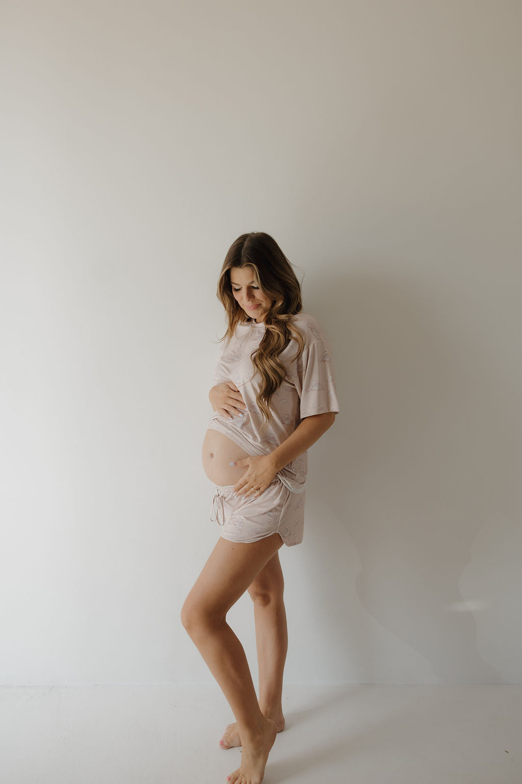 A pregnant woman with long wavy hair stands barefoot against a plain light wall, cradling her baby bump and looking down. She wears the Kendy 3 Women's Bamboo Short Set | Ruby—loose, breathable, hypo-allergenic bamboo top and shorts.