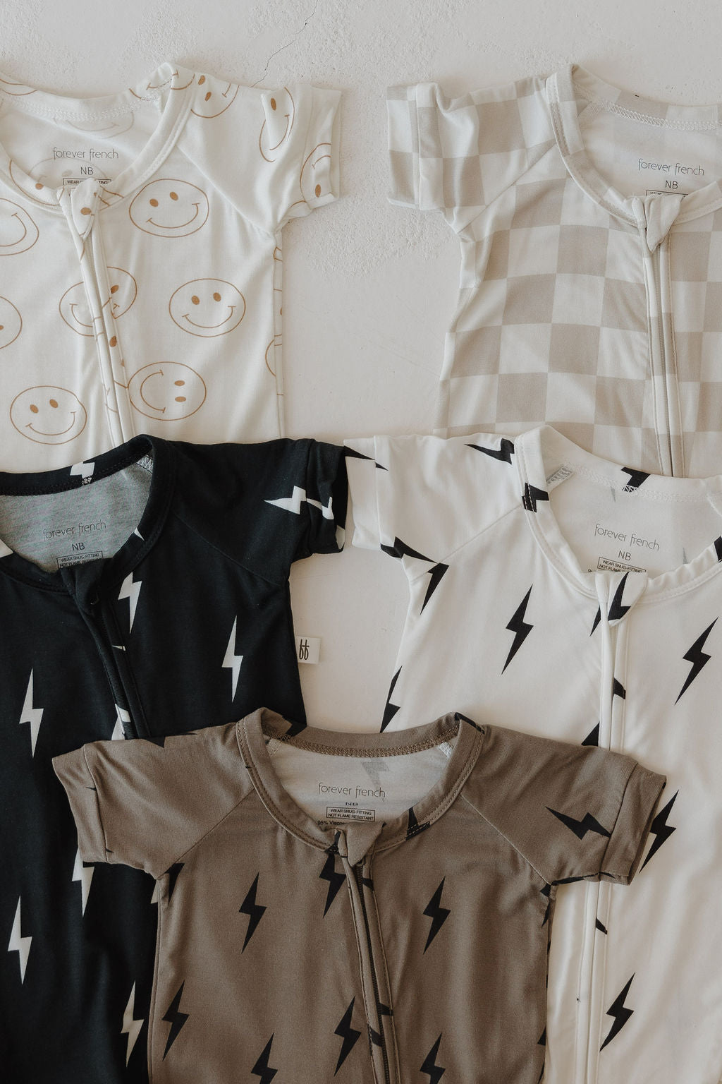Five forever french baby Bamboo Zip Shorties are laid flat, featuring designs like cream smiley faces, a checkerboard print, black with white lightning bolts, taupe with black lightning bolts, and brown & black lightning bolts.