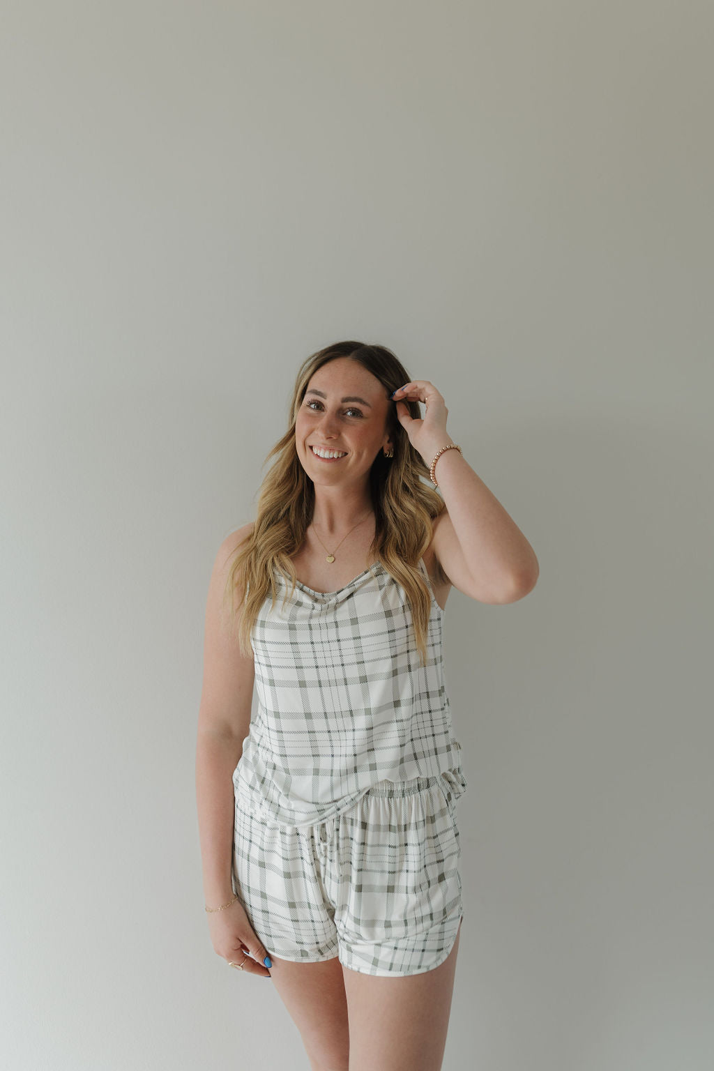 A woman with long hair, smiling against a plain background, showcases the elegance of her Cami Women's Bamboo Set in Picnic Plaid by forever french baby. Crafted from breathable fabric for comfort, she playfully holds one hand near her head, highlighting the chic sleeveless top and matching shorts.