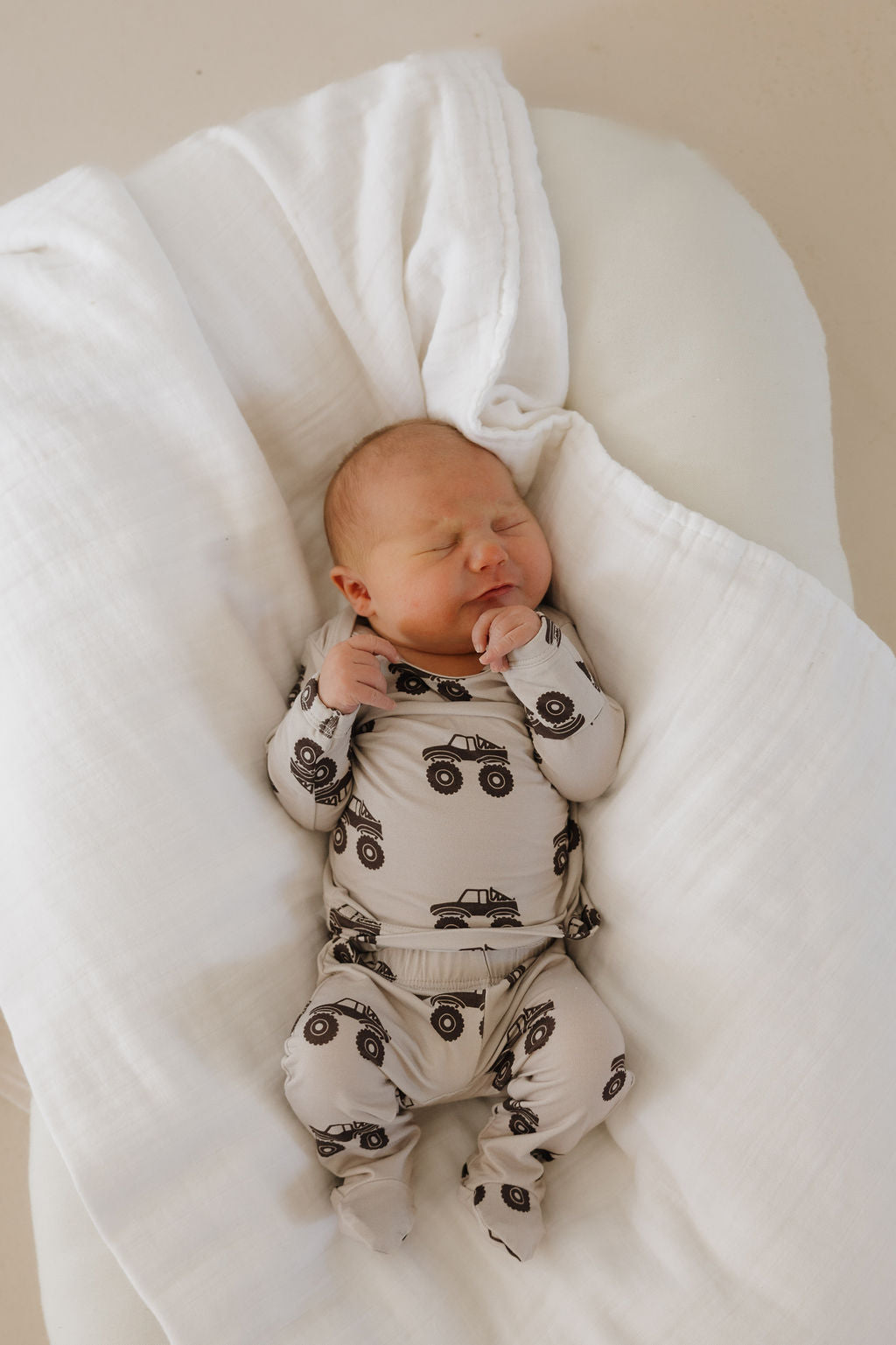 A newborn sleeps peacefully on a white blanket, wrapped in the cream-colored Sleepy Time Set | Monster Truck by forever french baby, featuring black monster truck prints.