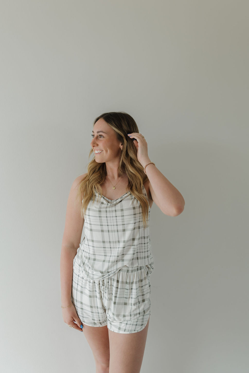 A person with long hair smiles and looks to the side, wearing the Cami Women's Bamboo Set in Picnic Plaid from forever french baby. The set includes a sleeveless plaid top and matching shorts made from breathable fabric, set against a plain, light-colored background.