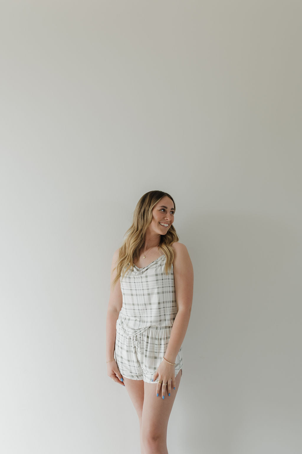 A woman smiles to her left, standing against a light backdrop in the Cami Women's Bamboo Set | Picnic Plaid by forever french baby. The sleeveless romper is made of breathable fabric. Her long hair falls in loose waves, and her arms are relaxed by her sides.