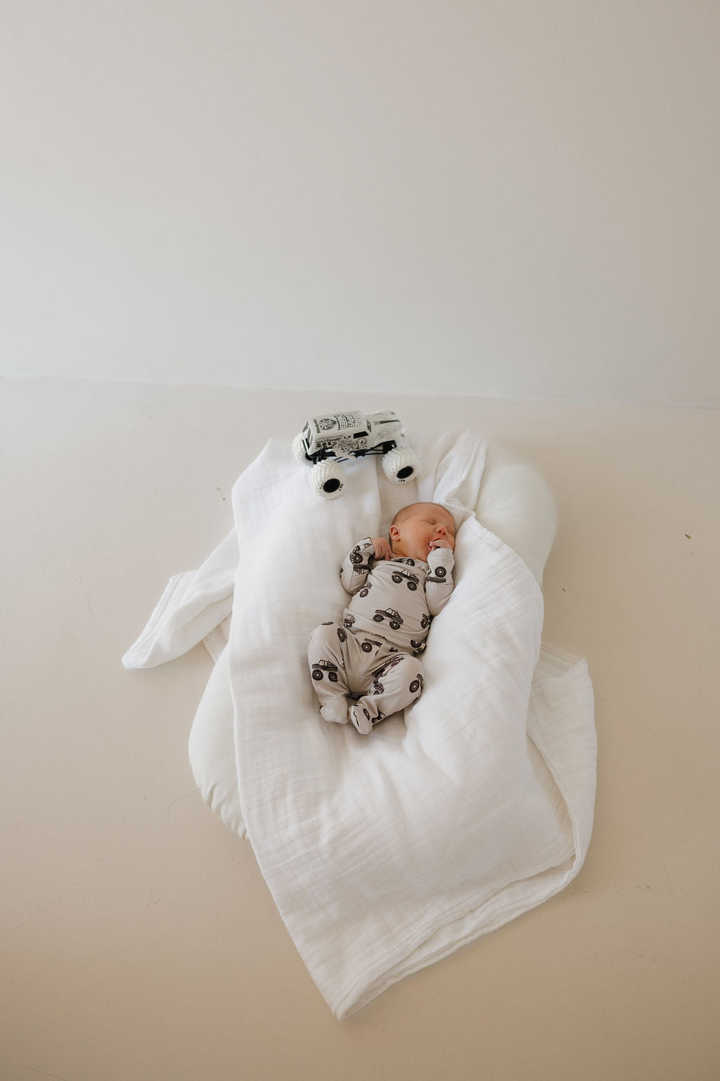 A newborn in the forever french baby Sleepy Time Set | Monster Truck onesie lies on a white blanket, partially wrapped, beside a small white toy car on a neutral-toned floor.