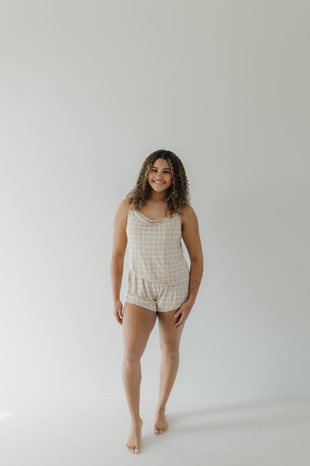 A person with curly hair is wearing the Cami Women's Bamboo Set | Grid by forever french baby, consisting of a sleeveless, checkered top and matching shorts. They are standing barefoot in a light-colored room with a neutral background, smiling at the camera.