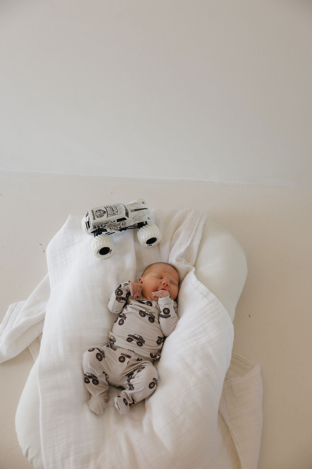 A baby in the hypo-allergenic white Sleepy Time Set | Monster Truck by forever french baby, featuring black monster truck prints, lies on a white blanket with a matching toy truck above—creating a minimalistic, peaceful scene.