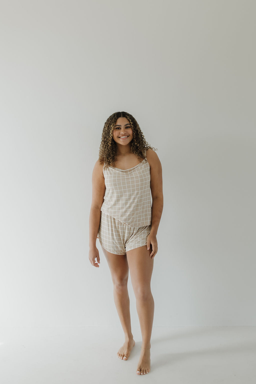 Barefoot against a white background, a person with curly hair wears the Cami Women's Bamboo Set | Grid by forever french baby. The breathable beige checkered pajama set includes comfy shorts and a sleeveless top as they smile and pose naturally.