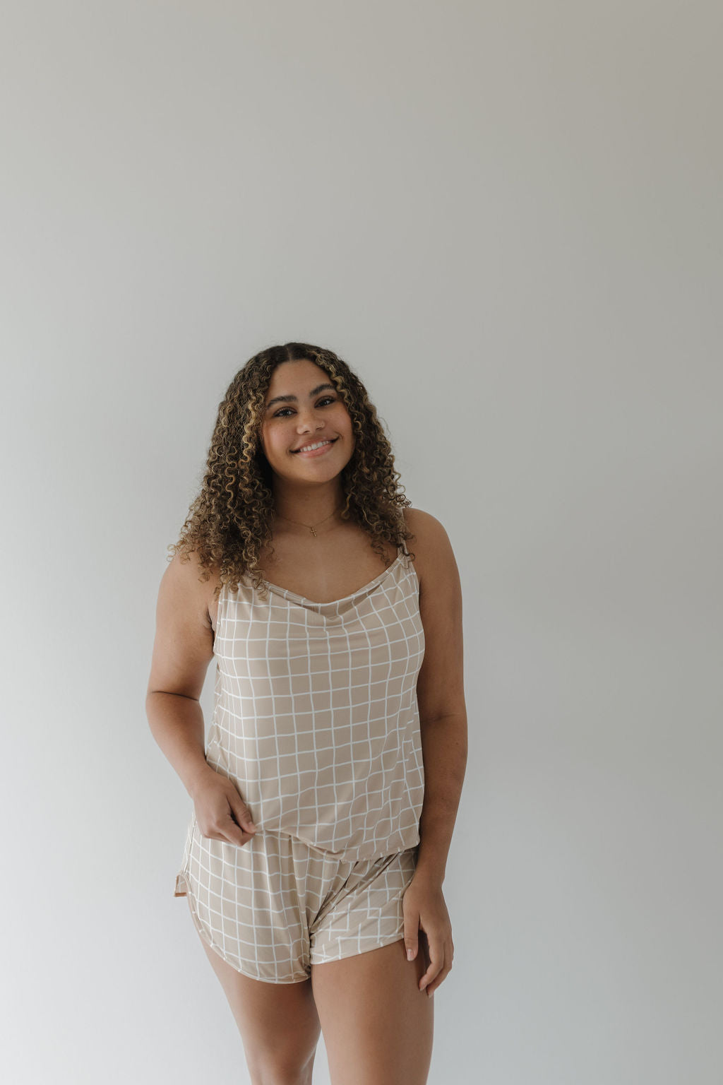 A woman with curly hair smiles at the camera wearing the Cami Women's Bamboo Set | Grid by forever french baby, a breathable and hypo-allergenic checkered tank top and shorts ensemble, against a plain light gray background.