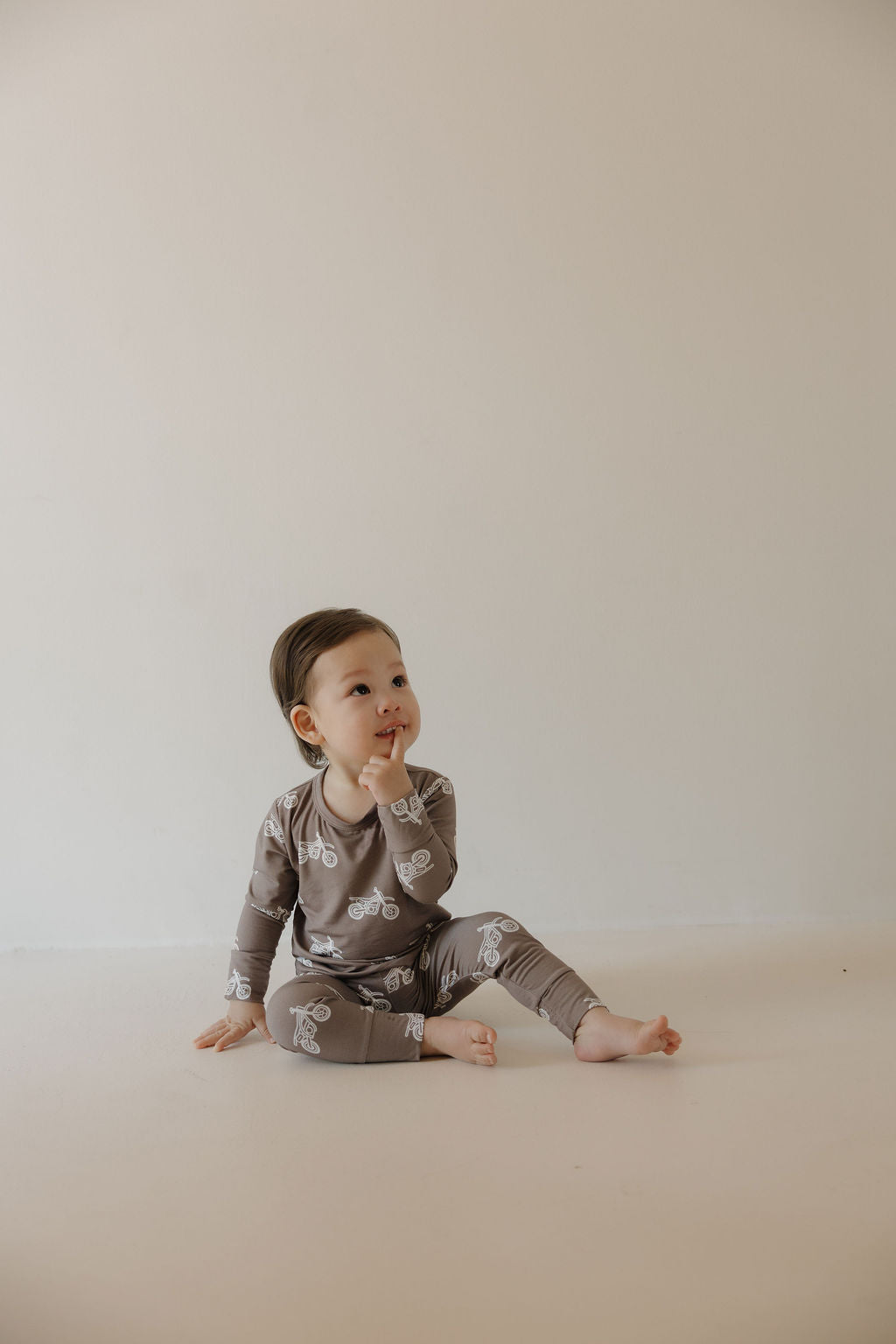 A young child sits on the floor against a beige background, dressed in the forever french baby Bamboo Footless Sleepy Time Set | Dirt Bike, looking up thoughtfully with one finger near their mouth.