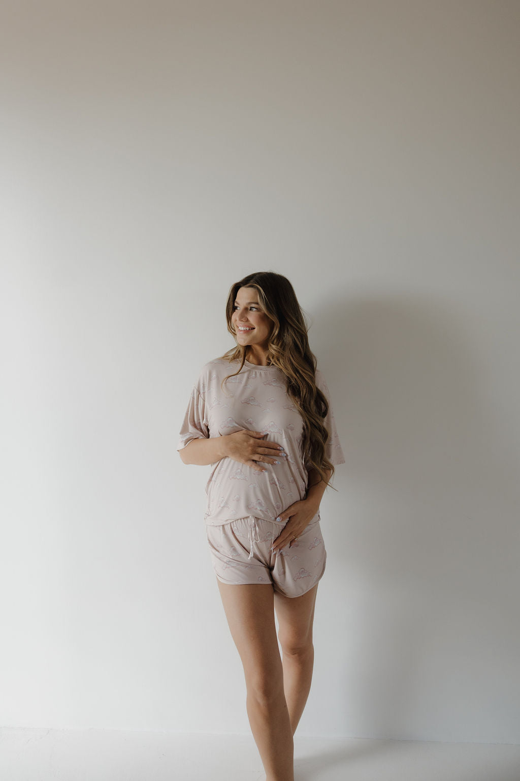 A woman with long hair smiles while standing against a light wall, gently cradling her belly. She wears the Kendy 3 Women's Bamboo Short Set in Ruby—light-colored, matching, and breathable pajamas.
