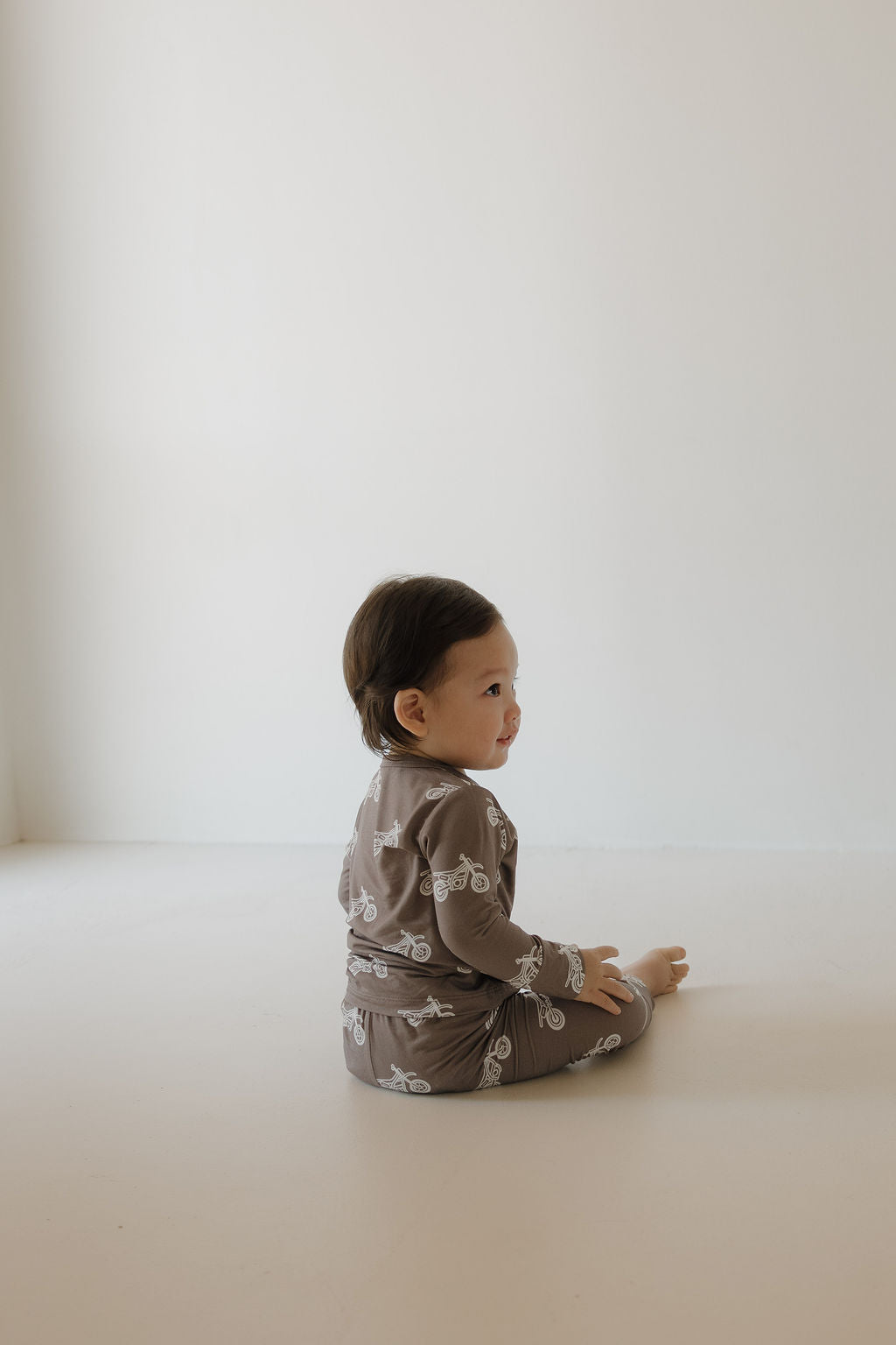 A toddler with short dark hair sits on a light floor, wearing the forever french baby Bamboo Footless Sleepy Time Set | Dirt Bike—brown hypo-allergenic pajamas featuring white bicycles. The plain background is softly lit, creating a cozy vibe.