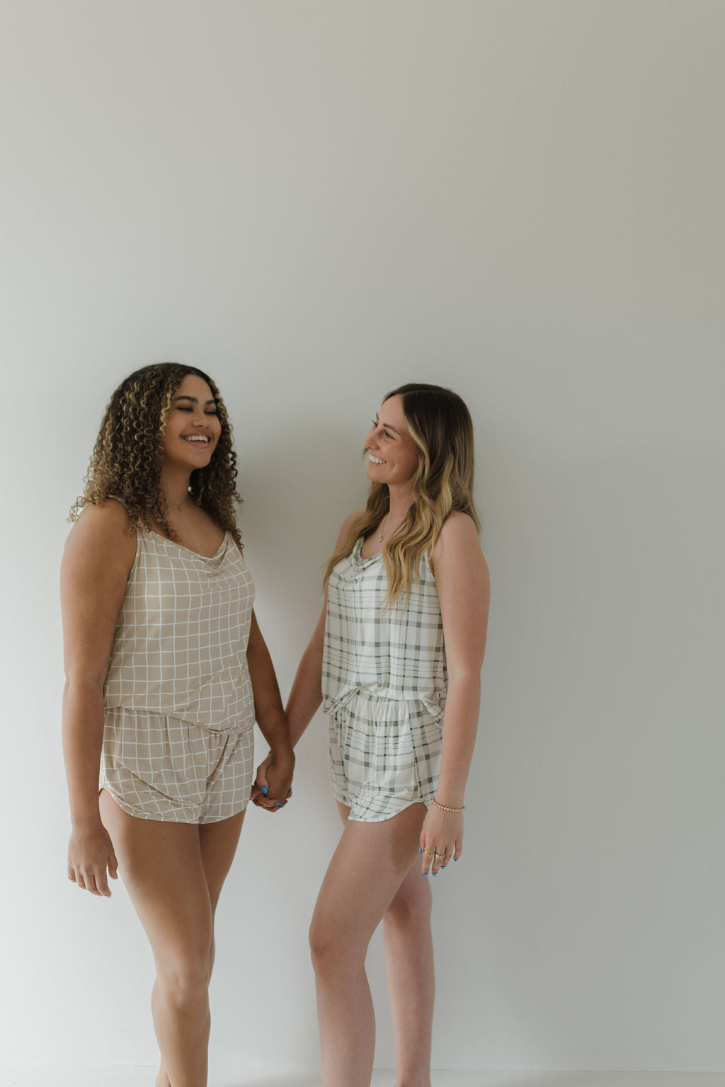 Two people hold hands and smile at each other in matching plaid outfits made of breathable material. One wears beige, the other white. They're wearing the Cami Women's Bamboo Set | Grid by forever french baby against a plain light-colored background.