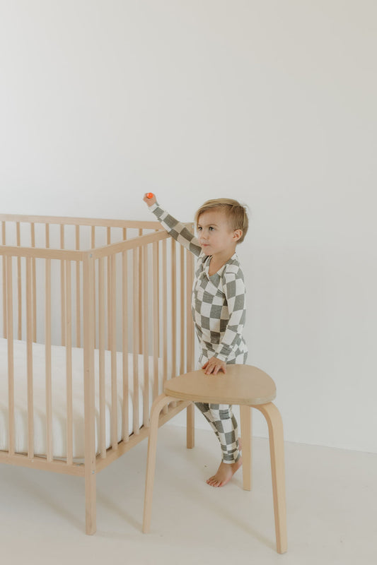 A child wearing forever french baby Bamboo Two Piece Pajamas in Oat & Sage stands on a wooden stool beside a light wood crib with a white mattress, reaching towards the wall. The pajamas' breathable fabric ensures comfort and ease, enhancing the serene ambiance.