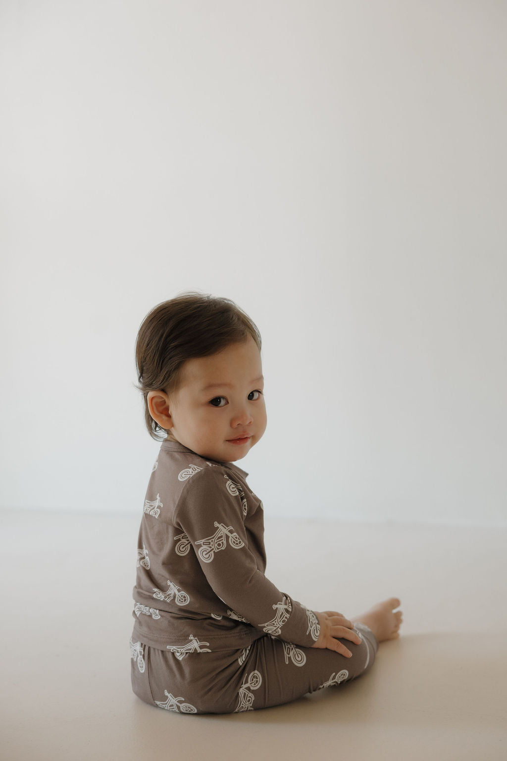 A young child with short dark hair sits on the floor, wearing the forever french baby Bamboo Footless Sleepy Time Set | Dirt Bike—brown pajamas with a white dirt bike pattern—looking back over their shoulder against a plain background.