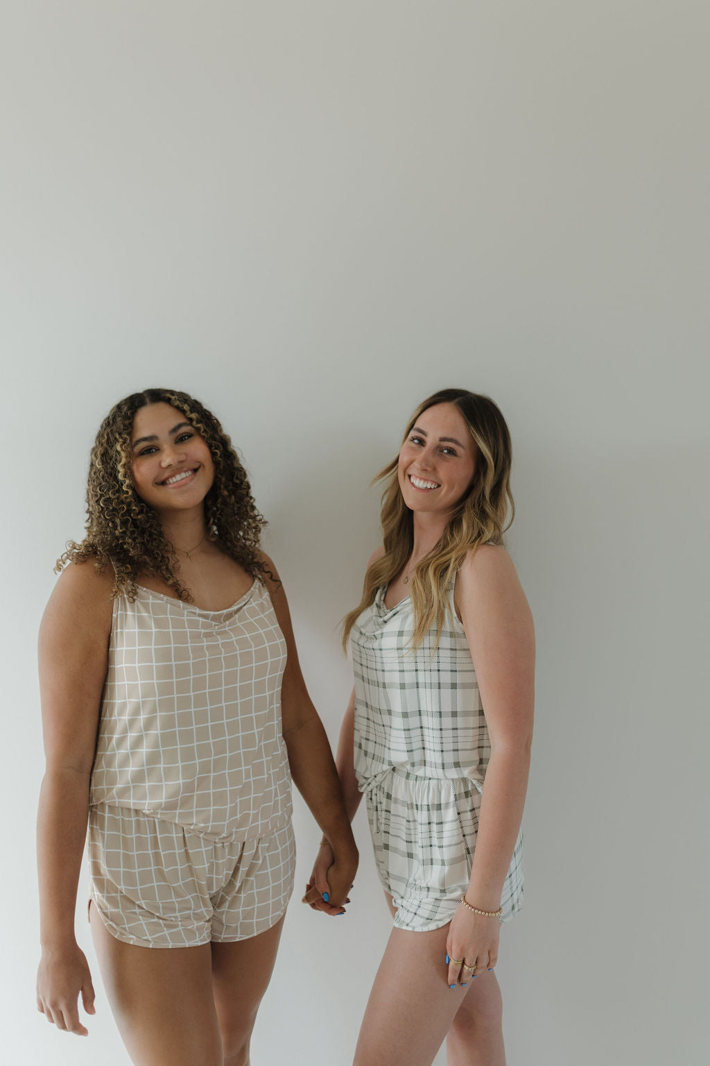 Two people hold hands and smile at the camera, wearing the Cami Women's Bamboo Set | Grid by forever french baby, featuring light-colored checkered rompers made from breathable bamboo fabric.