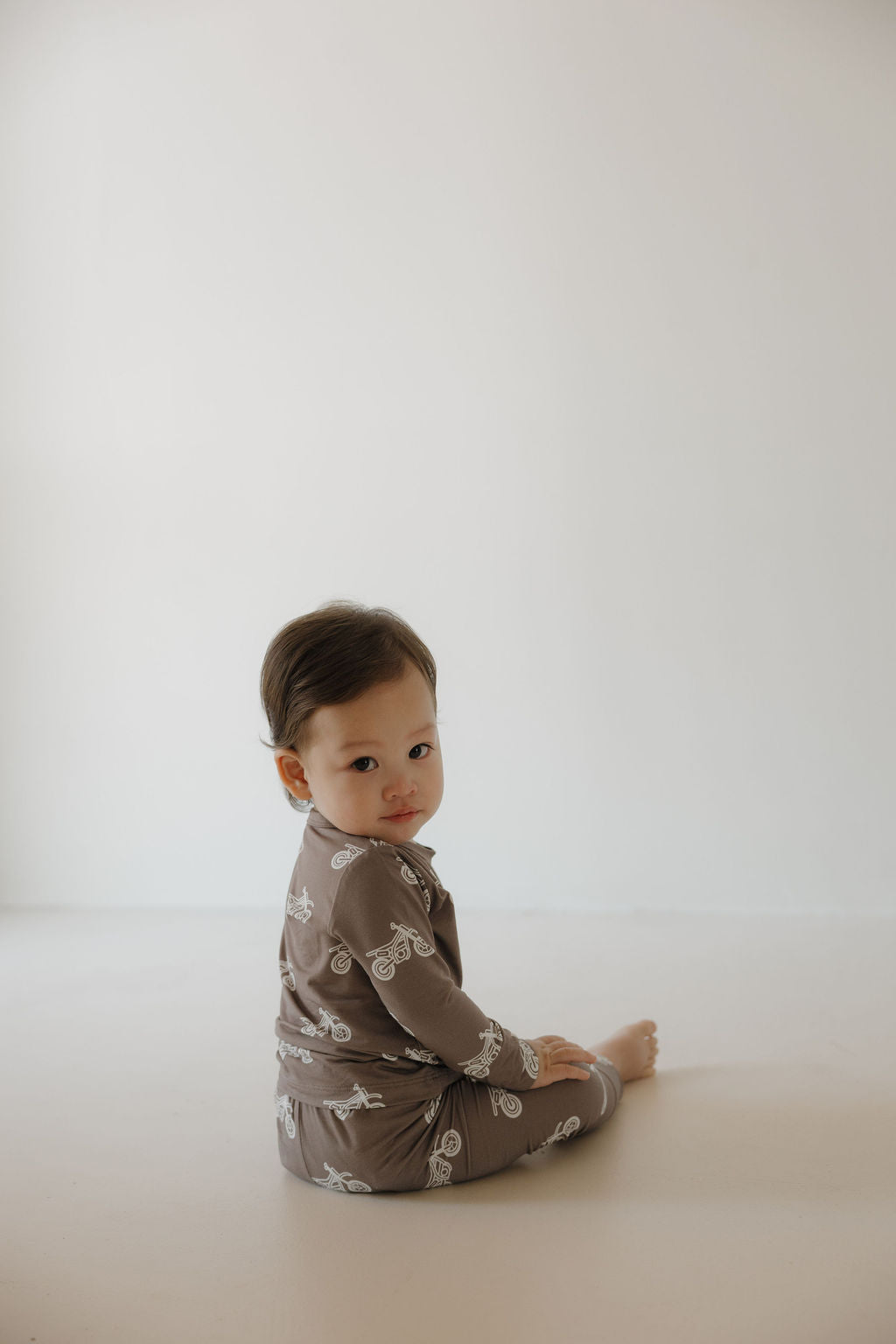 A young child with short brown hair sits on the floor wearing the forever french baby Bamboo Footless Sleepy Time Set | Dirt Bike, featuring brown pajamas with white bike patterns, looking over their shoulder at the camera against a light background.
