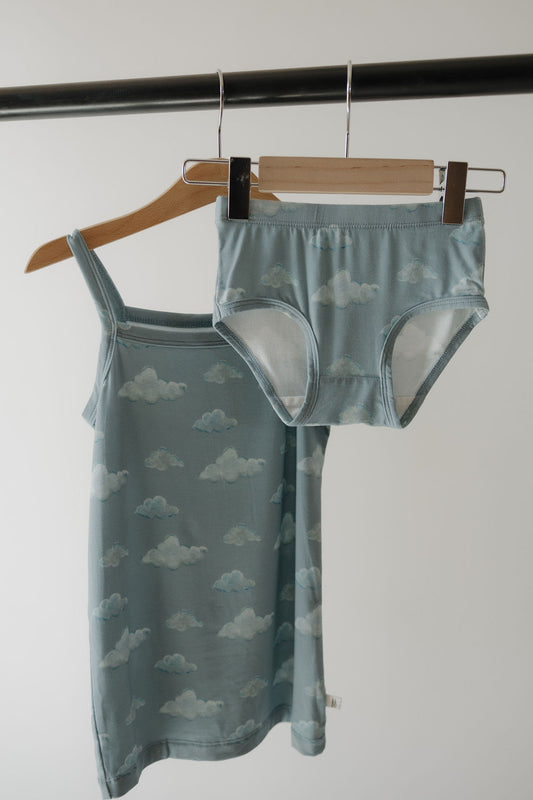 The Kendy 3 Toddler Bamboo Cami Set | Duke, featuring a light blue breathable camisole dress and matching underwear with a white cloud pattern, hangs on wooden and metal hangers against a plain background.