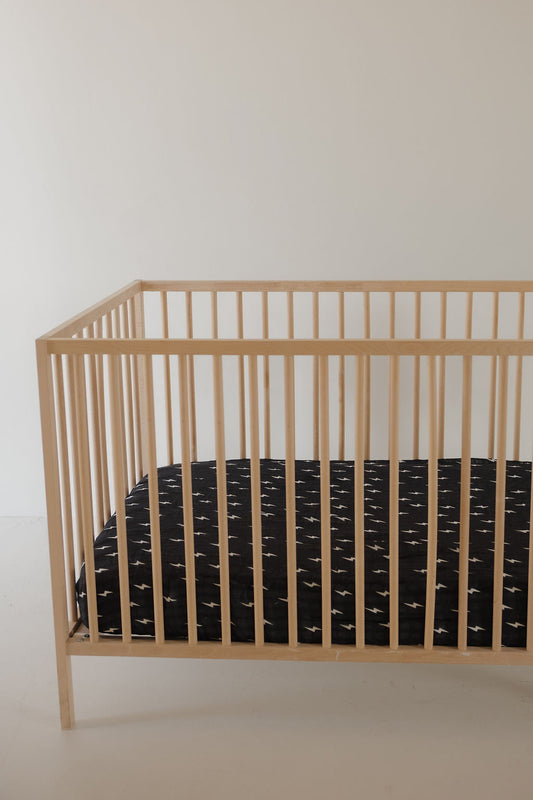 A minimalist wooden crib with a black mattress, topped with the forever french baby Muslin Crib Sheet | Midnight Bolt, sits against a plain light wall in an uncluttered room.