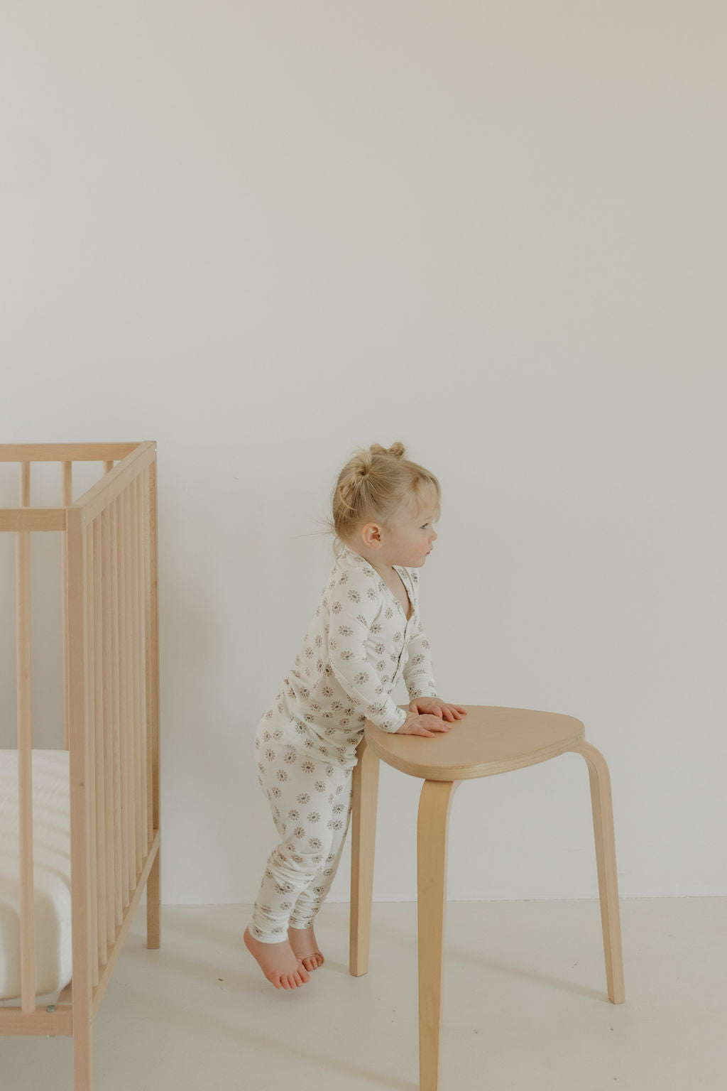 A toddler in forever french baby's Bamboo Two Piece Pajamas | Daisy Fields leans against a wooden stool, looking away. On the left is a wooden crib with a white blanket. The room features a minimalist style with light-colored floors and walls.