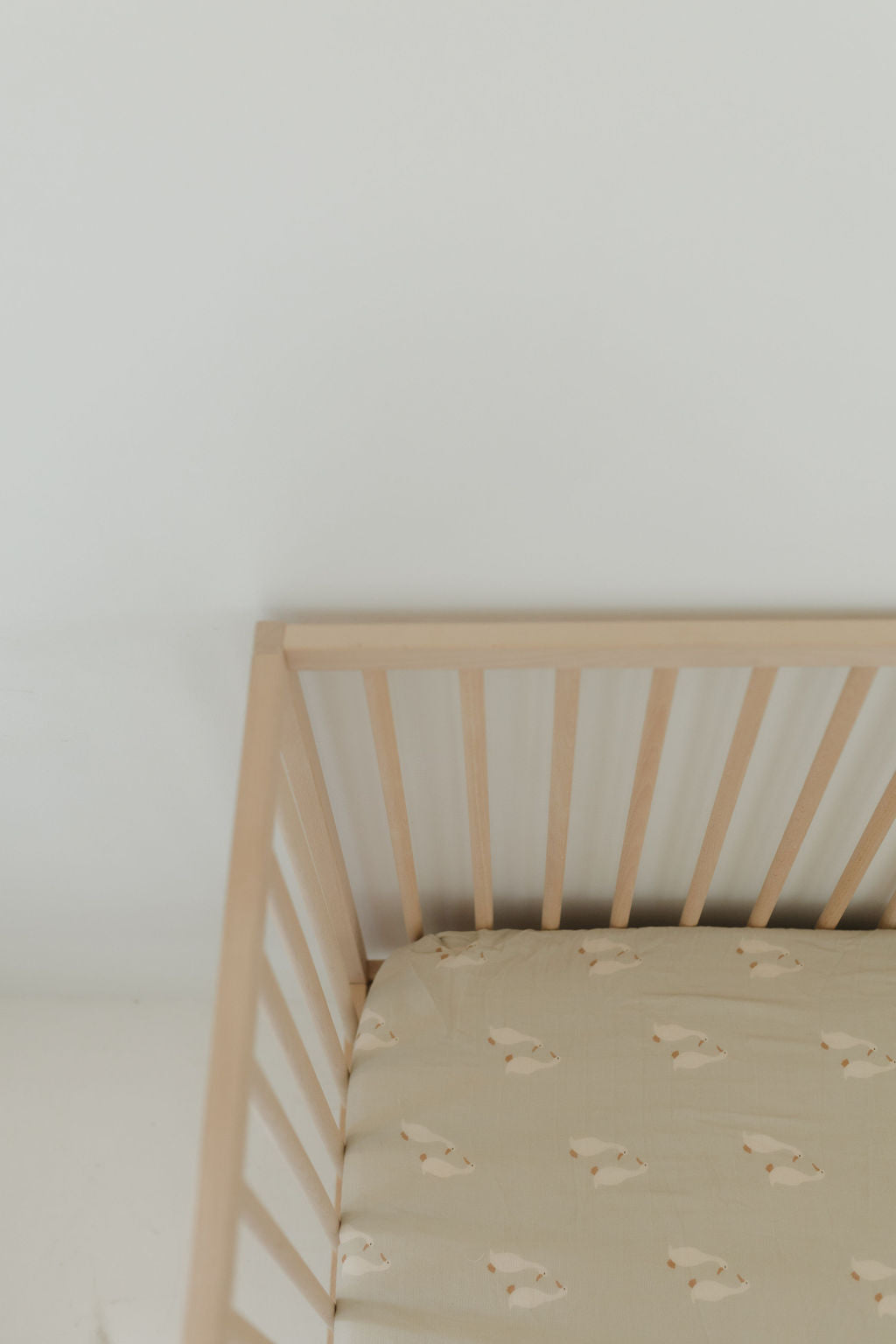 A minimalist wooden crib with light slats is paired with the Silly Goose muslin crib sheet by forever french baby, featuring a subtle animal print, all set against a plain white wall.
