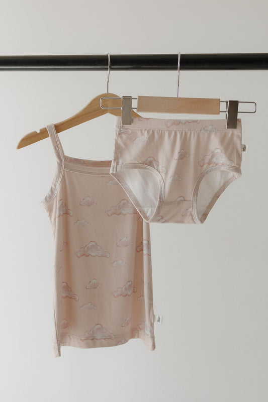 The Kendy 3 Toddler Bamboo Cami Set | Ruby, a pale pink tank top and matching underwear with white cloud patterns, made from breathable bamboo fabric, hangs on a black rack against a plain background.