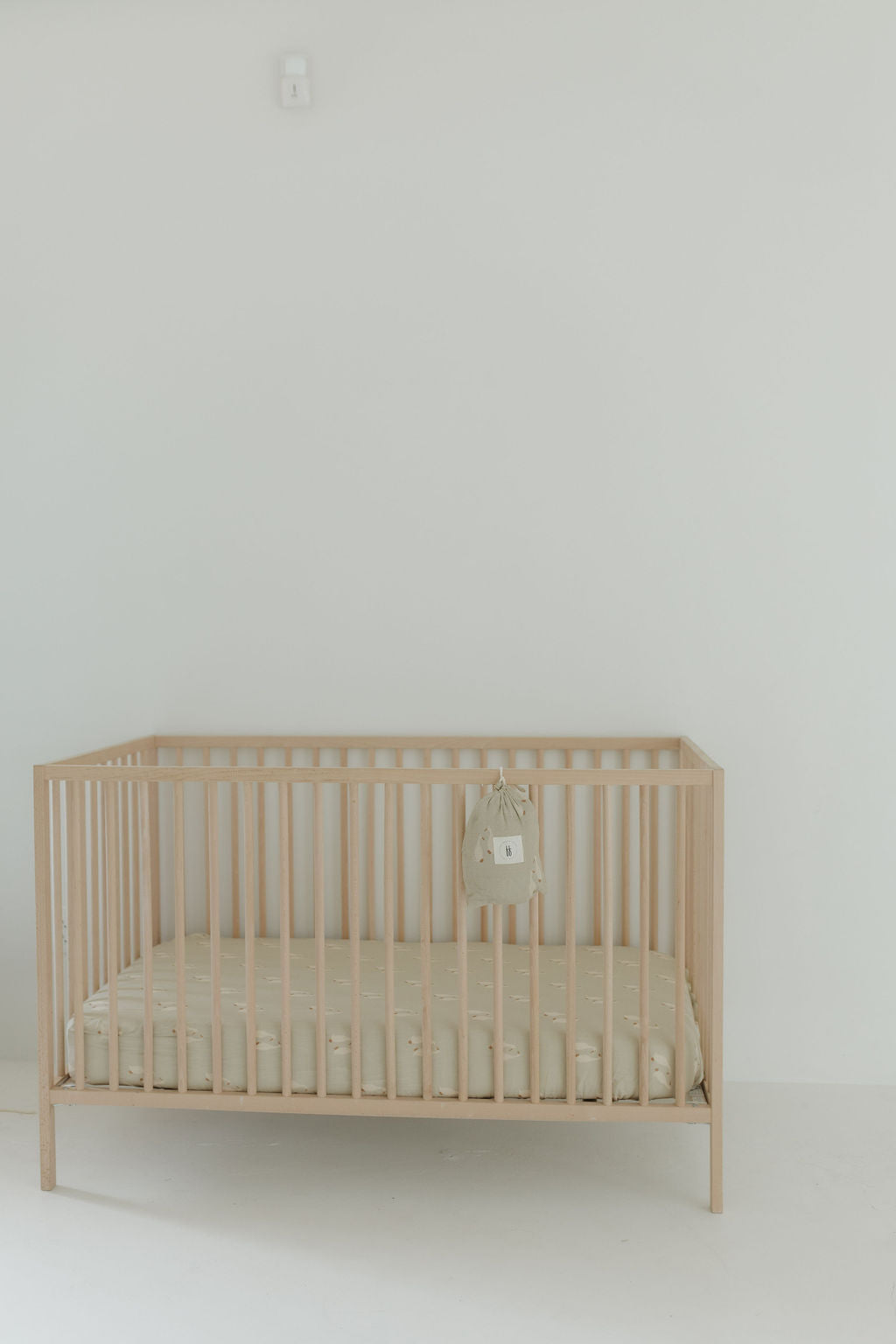 A minimalist nursery with a light wooden crib against a white wall, dressed in the Muslin Crib Sheet by forever french baby (Silly Goose). A small, light bag hangs on the side, completing the bright, uncluttered space.