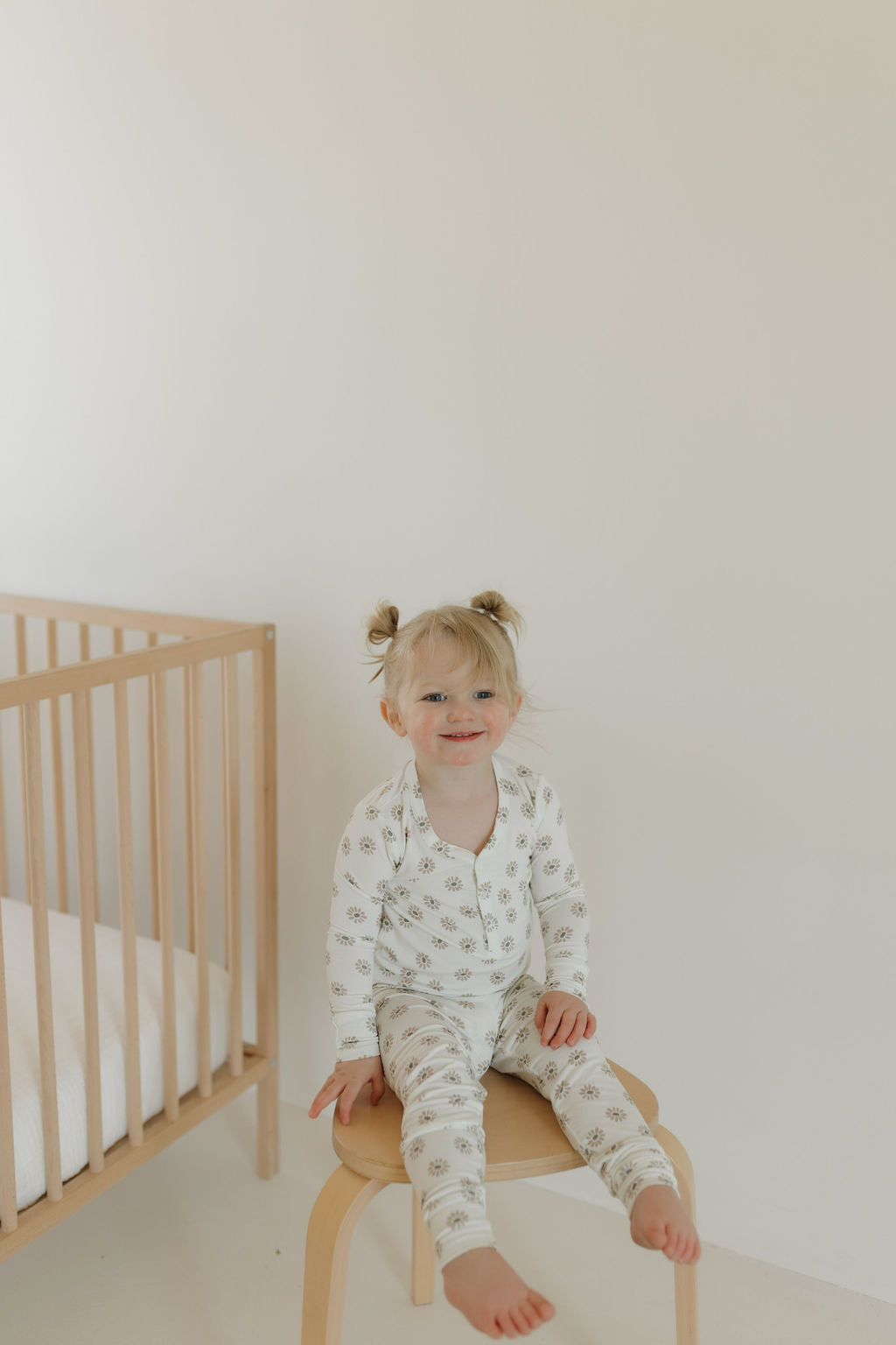 A smiling toddler with blonde hair sits on a wooden chair in a minimalist room, wearing forever french baby's Bamboo Two Piece Pajamas in Daisy Fields. A hypo-allergenic wooden crib is visible next to them against the plain white wall.