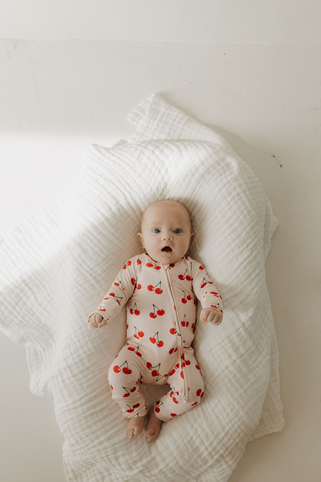 A baby lies on a white, textured blanket dressed in forever french baby's Bamboo Zip Pajamas | Cherry Orchard. Gazing upward with curiosity, the little one seems perfectly content in their hypo-allergenic clothing.
