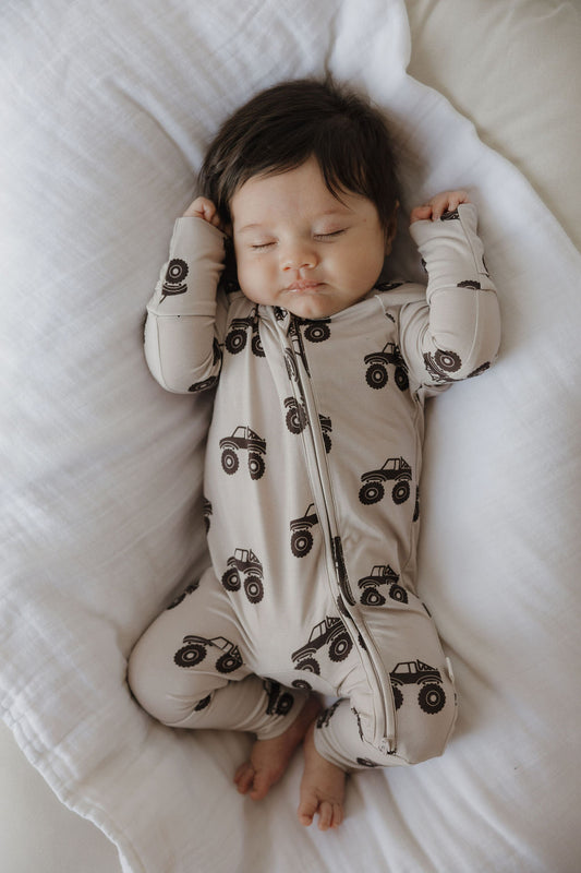 A baby with dark hair sleeps on its back on a white pillow, wearing forever french baby's Bamboo Zip Pajamas | Monster Truck in beige with black truck prints. The baby's arms are near its head and its legs are slightly bent.