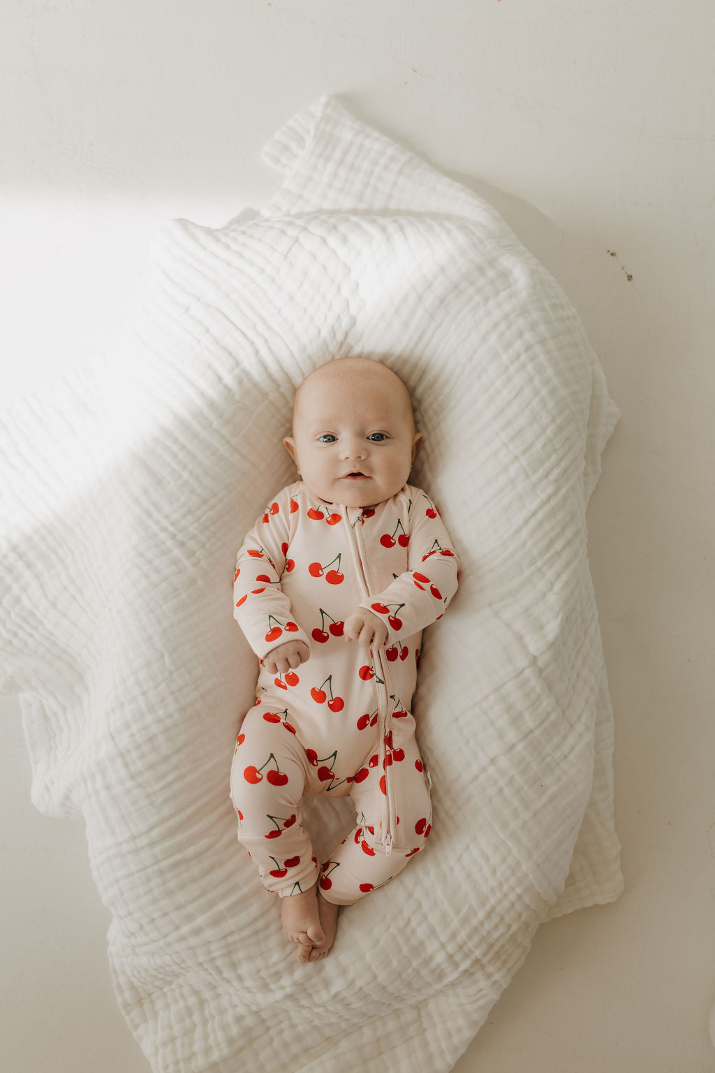 A baby lies on a soft white blanket wearing forever french baby's Bamboo Zip Pajamas in Cherry Orchard design. These breathable pajamas ensure comfort as the baby gazes upward with a neutral expression, surrounded by gentle natural light.