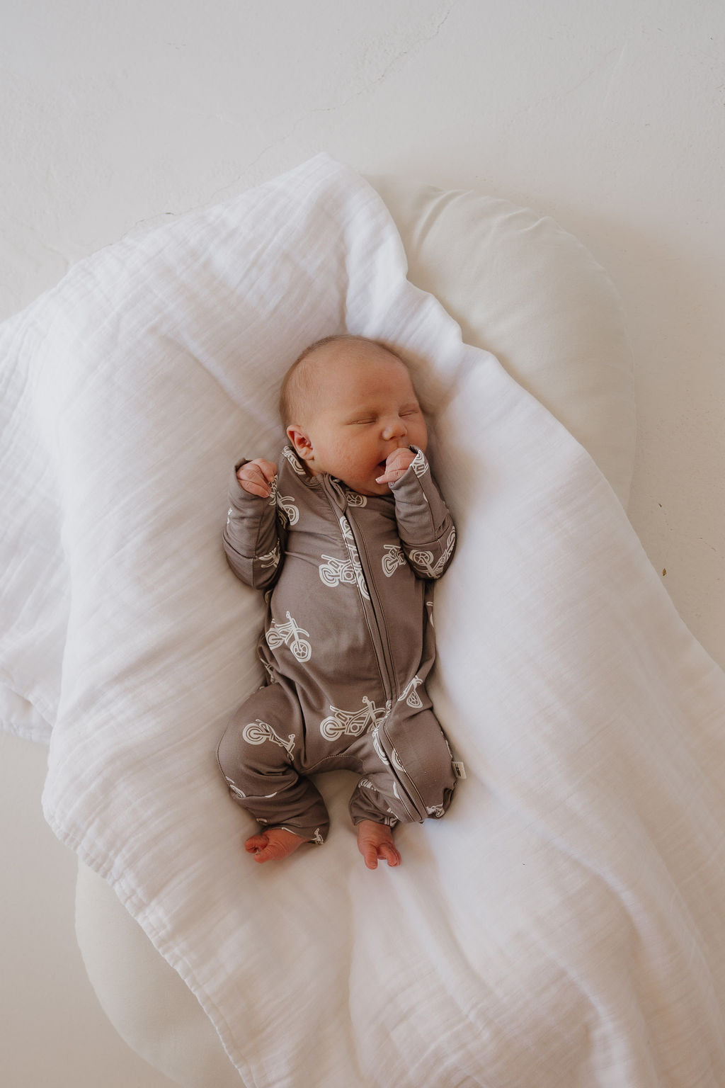 A newborn sleeps on a white, cushioned surface under a soft white blanket, wearing forever french baby’s Bamboo Zip Pajamas in the Dirt Bike pattern with one hand near their mouth.