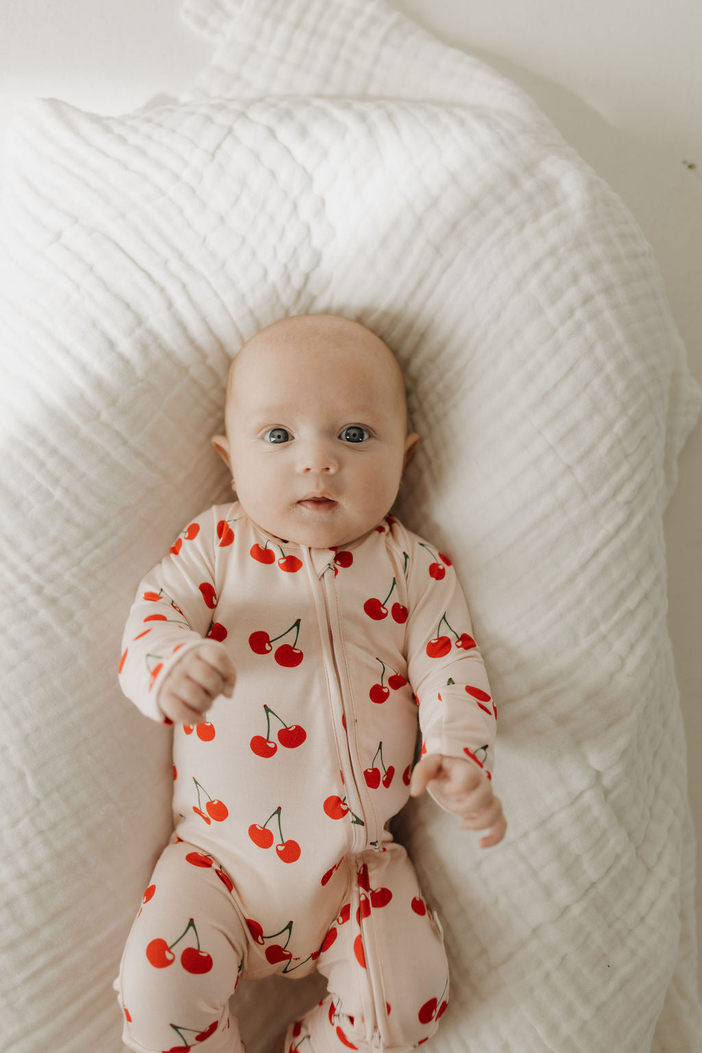 A baby with wide eyes and short hair lies on a white quilted blanket, comfortably wearing the "Bamboo Zip Pajamas | Cherry Orchard" by forever french baby—a charming, breathable, hypoallergenic onesie.