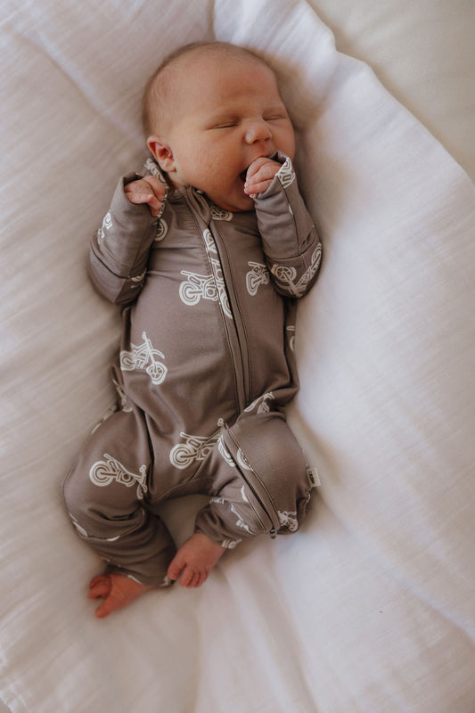 A newborn sleeps on a white blanket, wearing forever french baby Bamboo Zip Pajamas in the Dirt Bike print—one hand near their mouth, the other by their side.