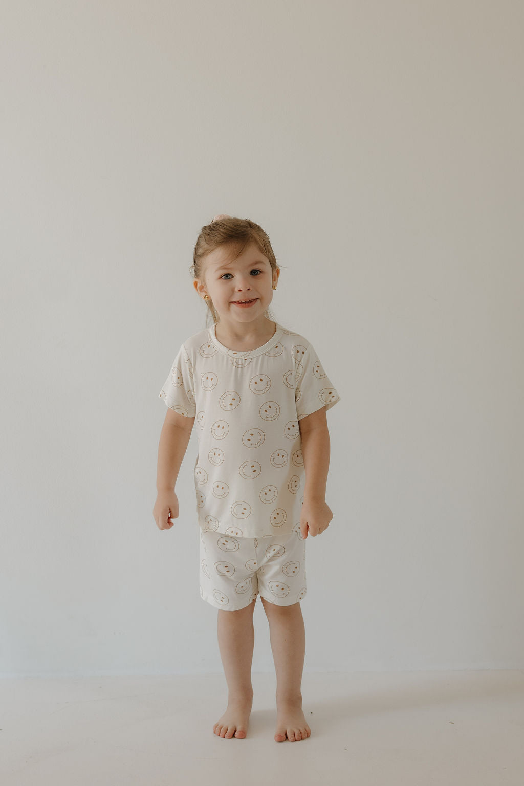 A young child with light brown hair smiles while wearing the forever french baby Bamboo Short Set | Just Smile, featuring matching bamboo pajamas and shorts with smiling faces, standing barefoot against a light-colored wall.