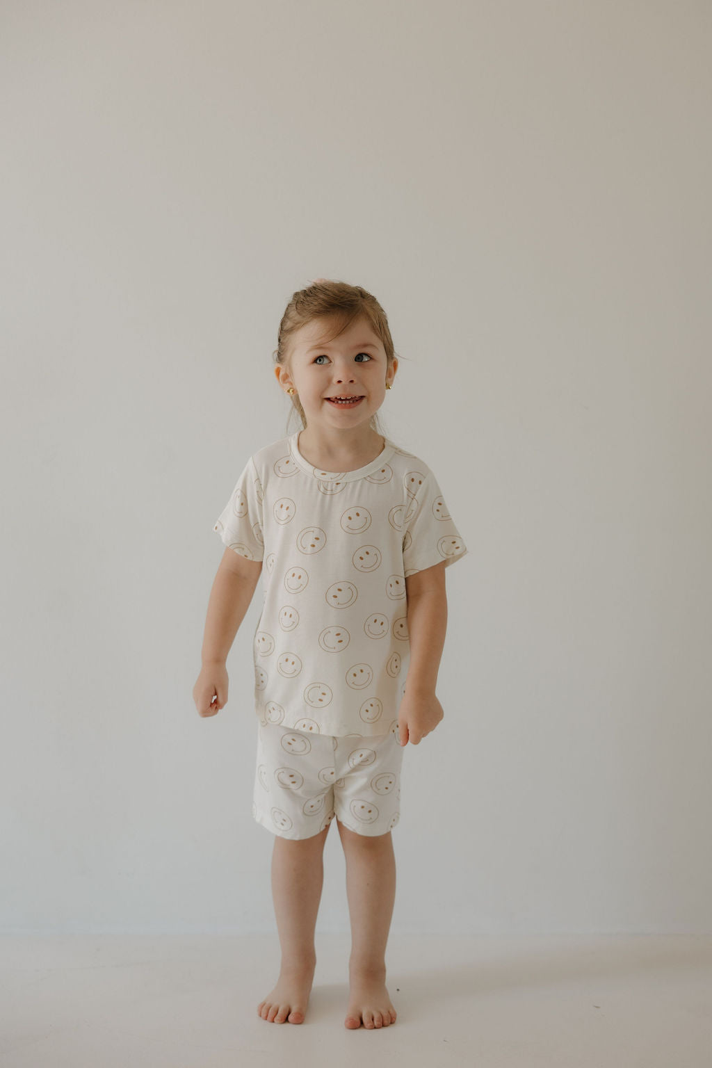 A young child with light brown hair smiles slightly, wearing the forever french baby Bamboo Short Set | Just Smile—cream-colored bamboo pajamas with a smiley face pattern—standing barefoot against a plain background. The breathable set keeps them comfortable.