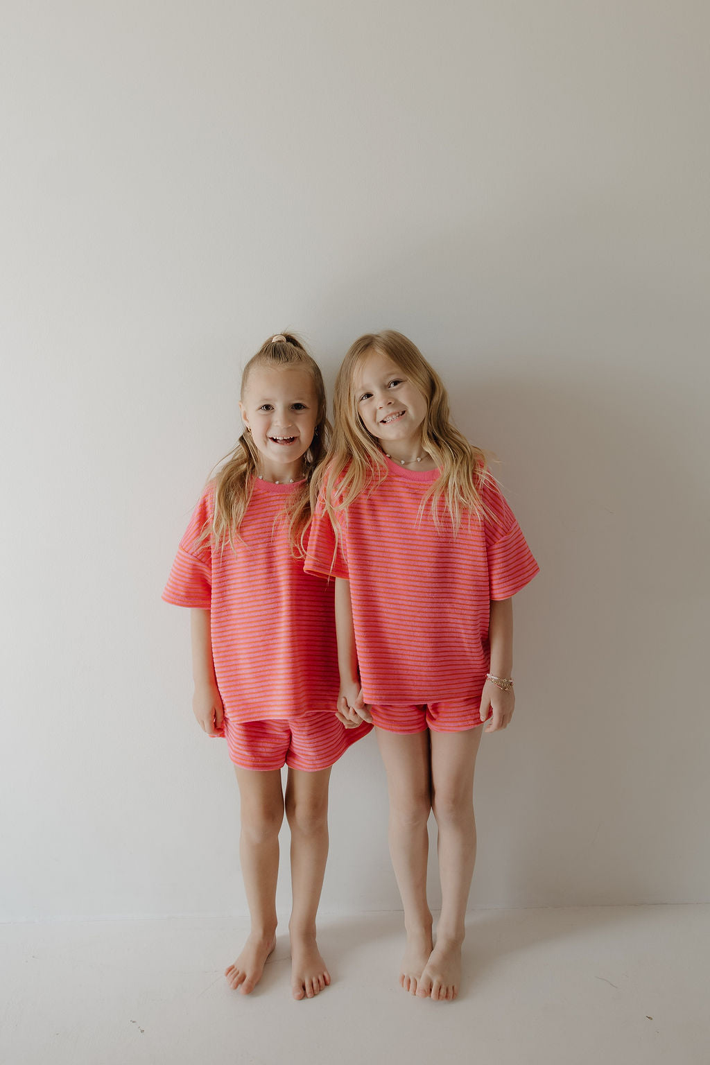 Two young girls wearing matching forever french baby Child Terry Short Set in Strawberry stand barefoot against a white wall, smiling and holding hands—capturing joyful family moments.