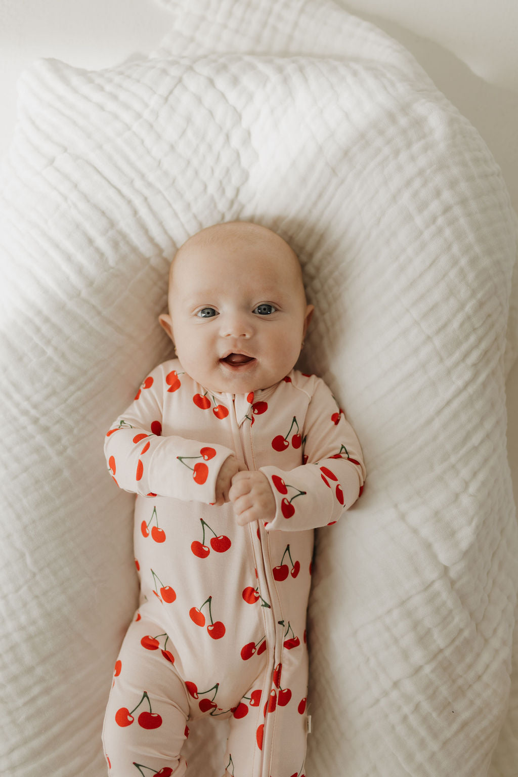 A baby lies on a soft, textured, white blanket wearing breathable, hypo-allergenic Bamboo Zip Pajamas with red cherry patterns by forever french baby. The little one looks up cheerfully, hands near the chest.
