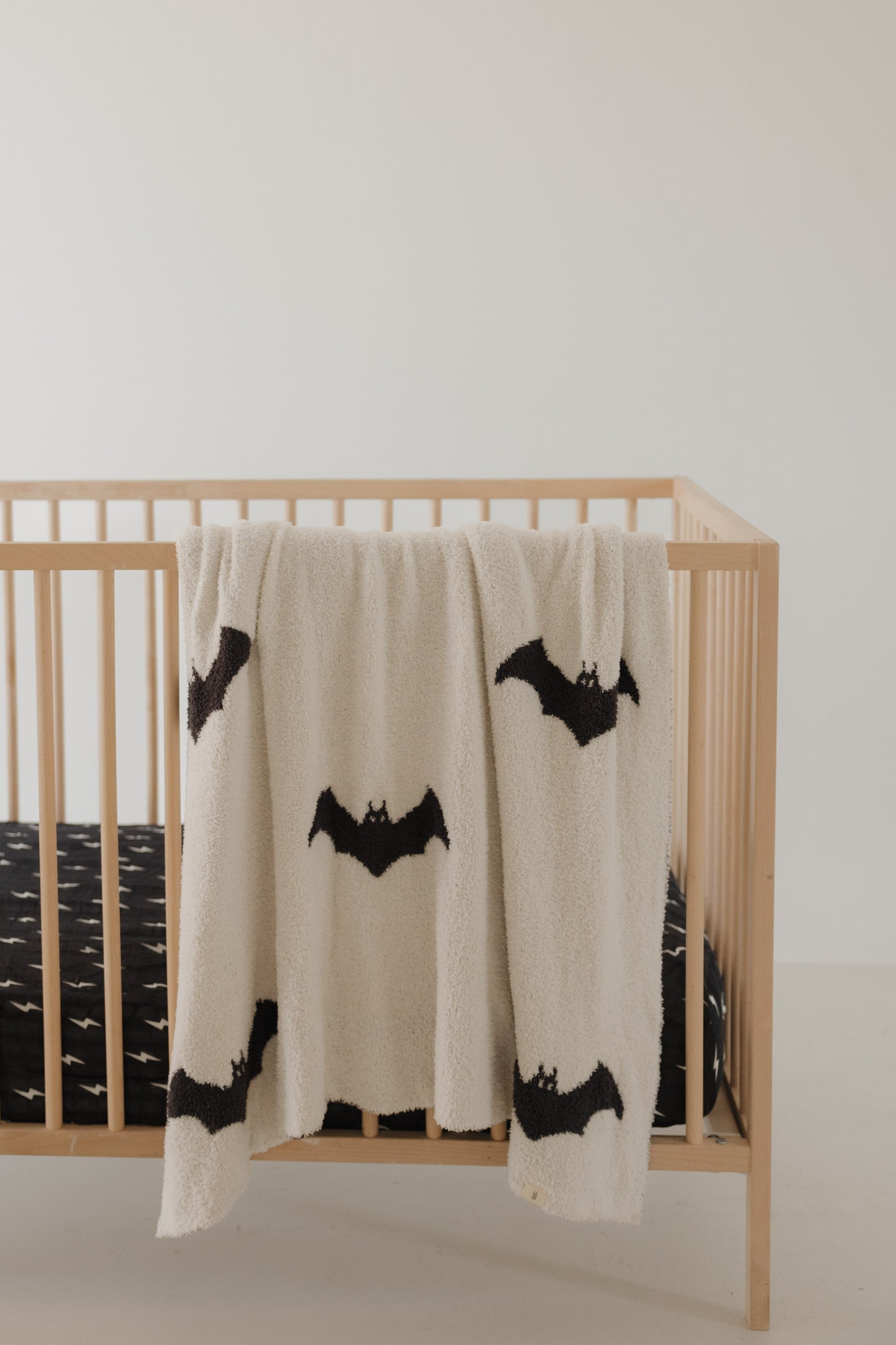 A wooden crib with a black mattress showcases the Plush Blanket | It’s Bats! by forever french baby, draped over the side. Featuring large black bat designs and crafted from Oeko-Tex certified materials, this soft baby blanket adds a touch of comfort to the minimalistic setting.
