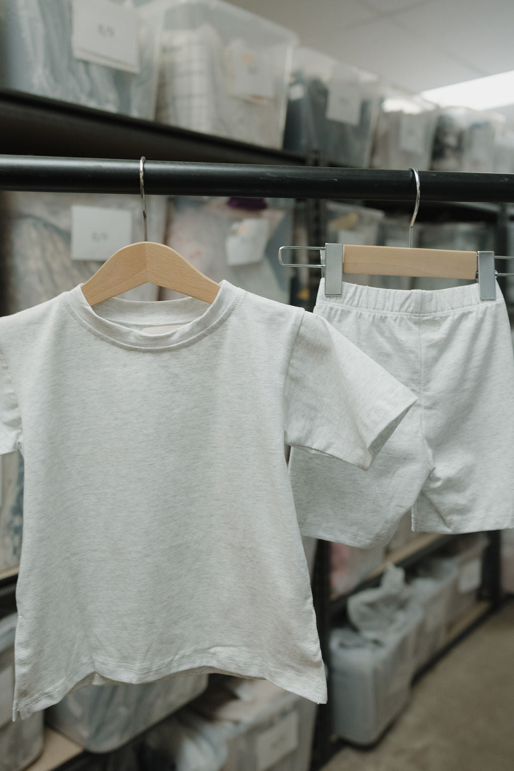 The forever french baby Child Short Set | Heather, with an oversized tee and biker shorts, hangs on wooden hangers from a clothing rack in a storage room filled with labeled plastic bins.