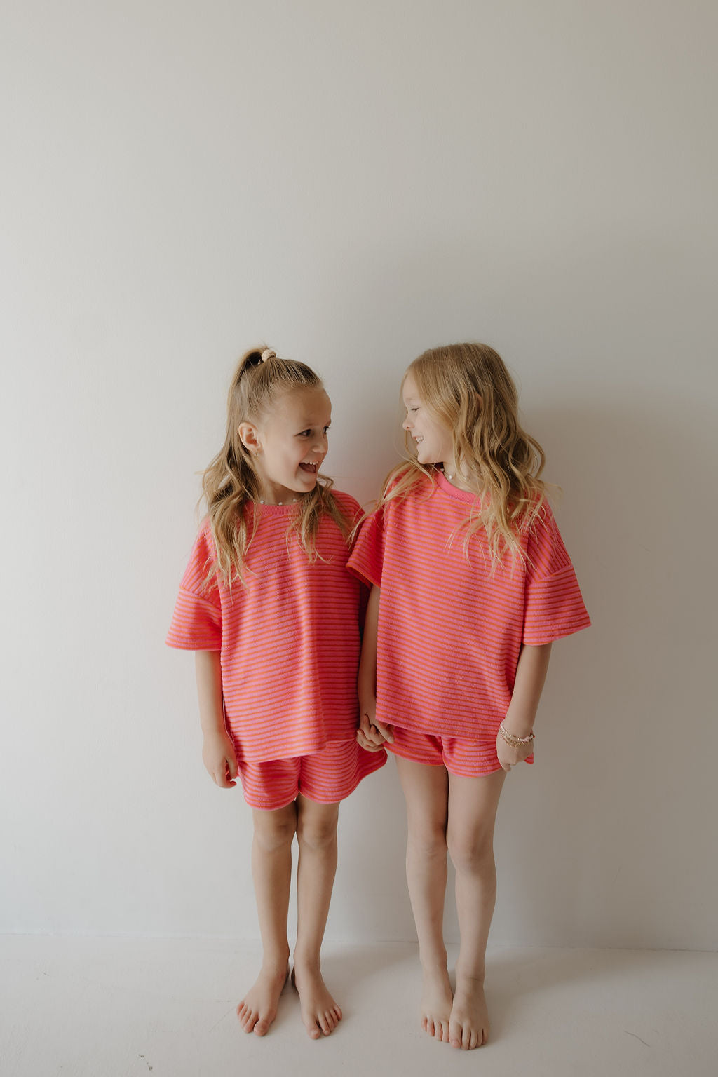 Two young girls stand barefoot against a white wall, smiling at each other in matching forever french baby Child Terry Short Sets | Strawberry, capturing the joy of simple family moments in coordinating pink striped outfits.