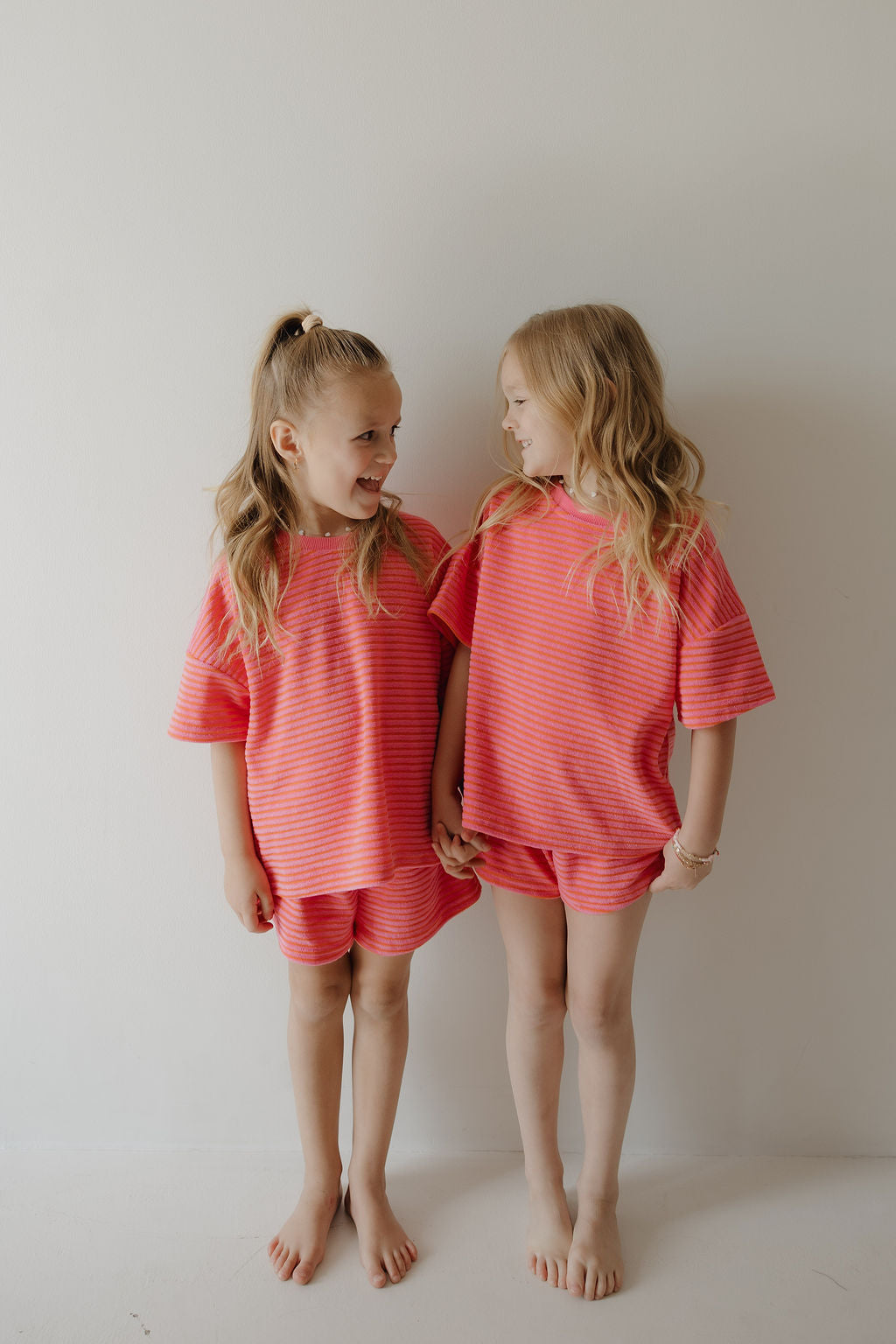 Two young girls with long blonde hair stand barefoot against a white wall, holding hands and smiling at each other in matching forever french baby Child Terry Short Set | Strawberry outfits—capturing a perfect family moment.