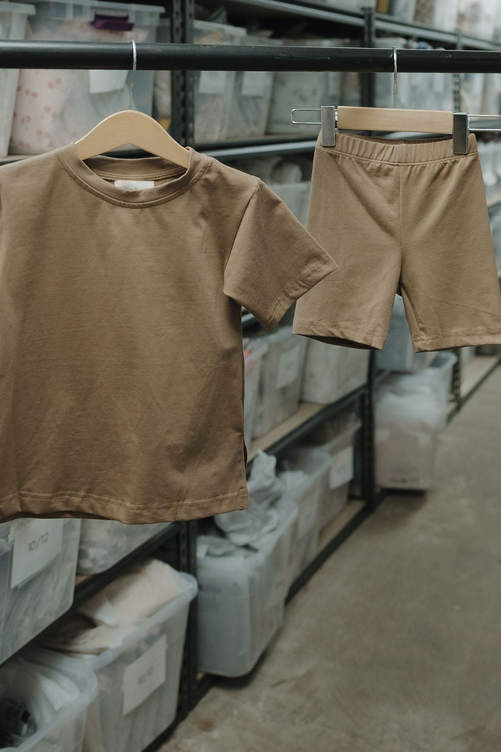 The "Child Short Set | Patina" by forever french baby, a beige oversized tee and biker shorts set, hangs on a rack in a storage room filled with clear bins of folded clothes.