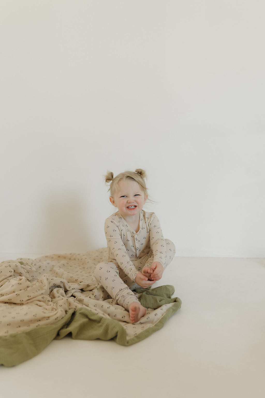 A young child with blonde hair in ponytails sits on a white floor, wearing a cream outfit with a subtle pattern, smiling and holding their feet beside the Muslin Quilt | Flora by forever french baby in beige and green hues.