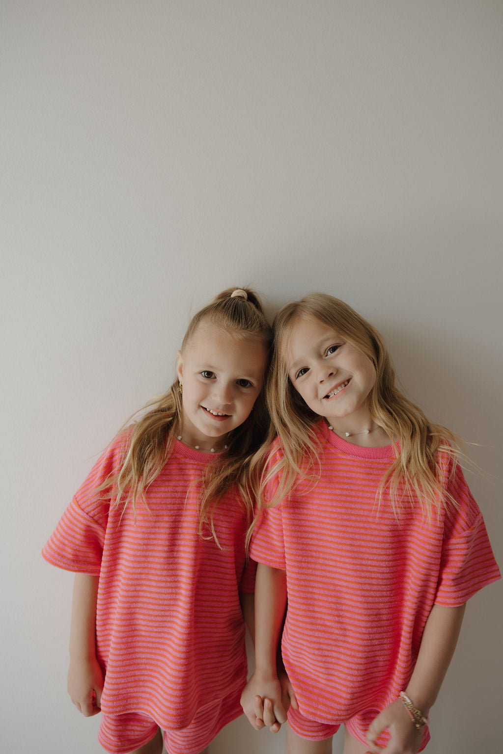 Two young girls with long blonde hair, wearing matching forever french baby Child Terry Short Set | Strawberry outfits, stand smiling and holding hands against a white wall, sharing a sweet family moment.
