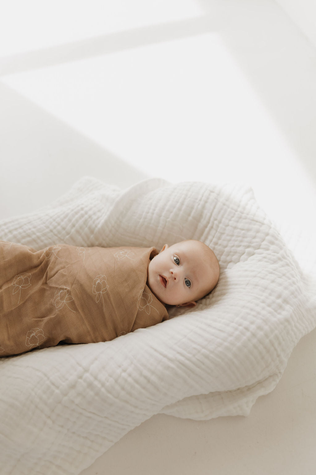 A baby wrapped in the forever french baby Muslin Swaddle | Sweet Dreams Floral lies peacefully on a white cushioned surface. Soft sunlight filters in, casting gentle shadows, creating a calm and serene atmosphere.