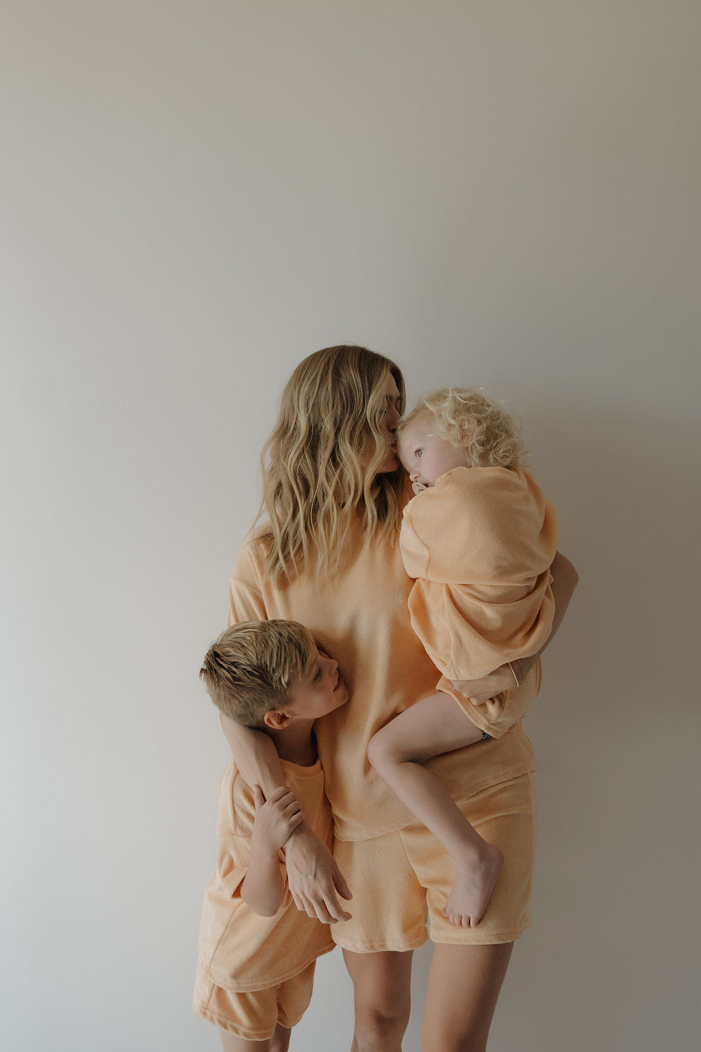 A woman in peach loungewear hugs two children against a plain background, capturing a heartfelt family moment. One child wears the forever french baby Child Terry Short Set in Sherbert and hugs her waist while the other rests on her shoulder.
