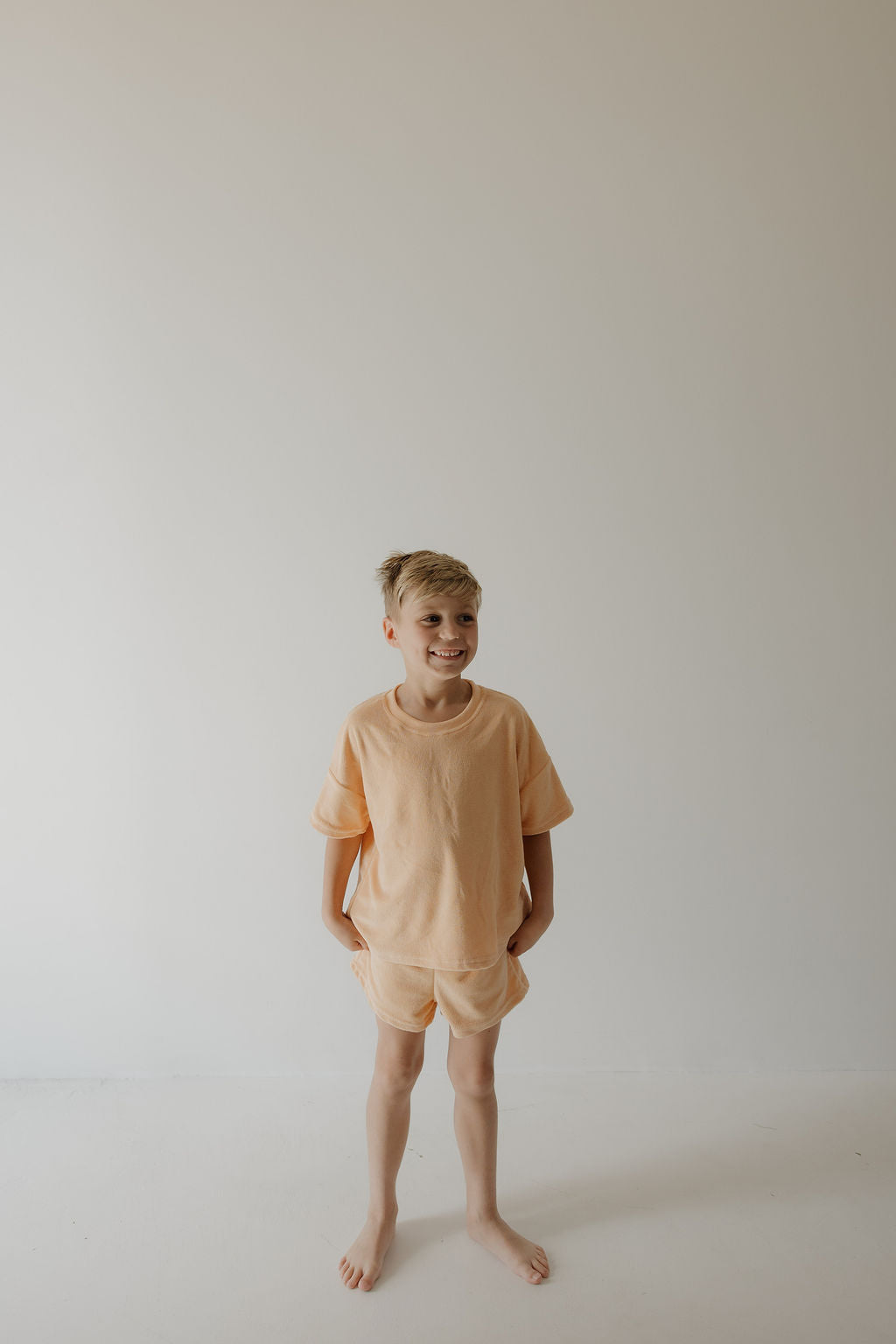 A young blond boy stands barefoot against a white wall, smiling in a forever french baby Child Terry Short Set | Sherbert, hands in his pockets—perfect for cozy family moments.