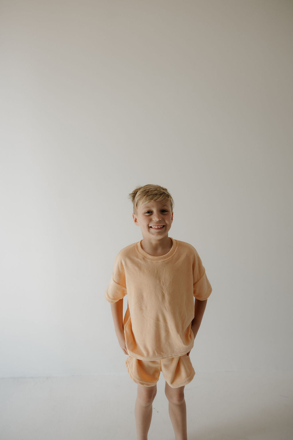 A young blonde boy smiles at the camera in the forever french baby Chid Terry Short Set | Sherbert, standing against a plain light background with his hands in his pockets—perfect for family moments.